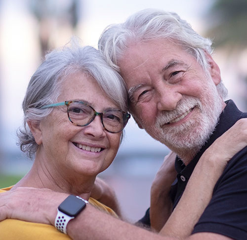 Elderly woman hugging her significant other over the shoulders and smiling at the camera.