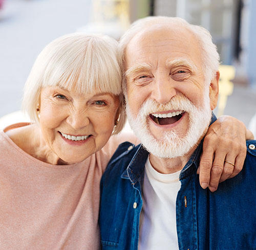 Elderly woman hugging her significant other over the shoulders and smiling at the camera.