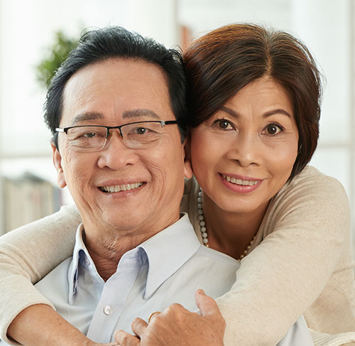 Elderly woman hugging her significant other over the shoulders and smiling at the camera.