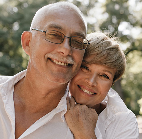 Elderly woman hugging her significant other over the shoulders and smiling at the camera.