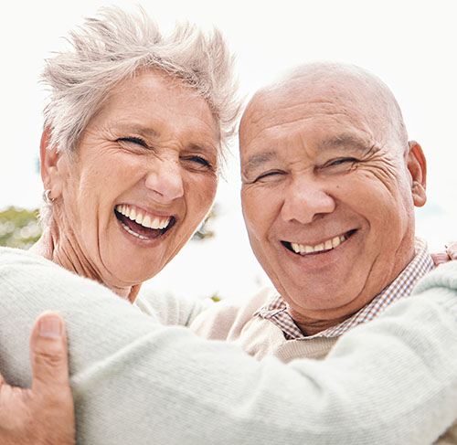 Elderly woman hugging her significant other over the shoulders and smiling at the camera.