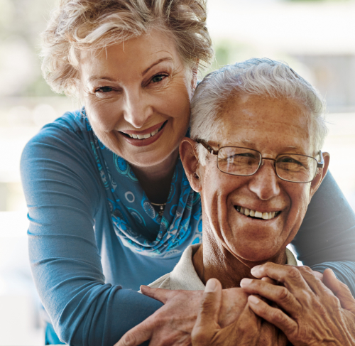 Elderly
                    woman hugging her significant other over the shoulders and smiling at the camera.